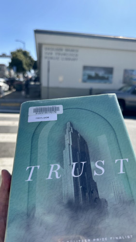 Cover of Hernán Díaz’s Trust in front of the blurry side of the Excelsior branch of the San Francisco public library