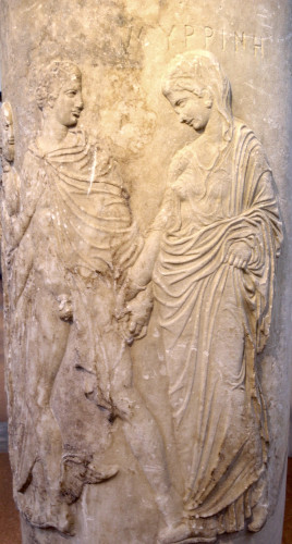 Relief of the god Hermes taking a dead woman's hand in a tender gesture on their way to the underworld. Hermes wears his winged boots and a traveller's cloak, otherwise he's naked. Myrrhine is veiled, wearing a long dress and a himation over it.