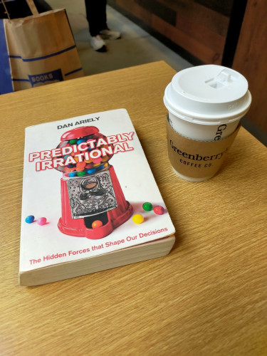 The photo is of a brown wooden table top. The paperback book is white with a red gumball machine on the cover. To the right is a white plastic-lidded paper coffee cup with a brown sleeve that says GREENBERRY COFFEE CO.