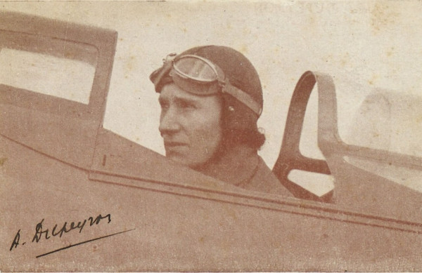 Andrée Dupeyron in sepia black and white photo. Seated in open cockpit of mid C20th plane wearing leather flying helmet and goggles pushed to forehead. in profile facing left. signature A Dupeyron bottom left