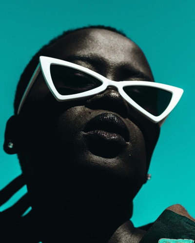 Striking headshot of a Black woman with a close-cropped haircut wearing oversized triangular-shaped white sunglasses. She is set against an aqua background.
