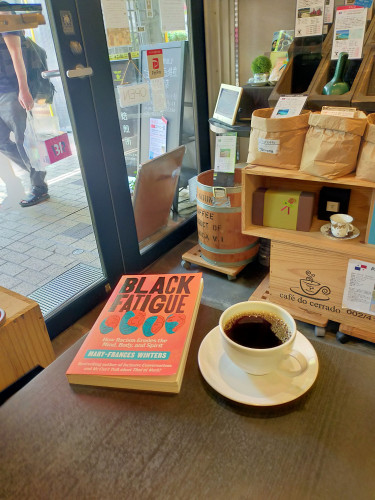 Photo is of the inside of the cafe. We can see brown bags of raw coffee beans on display in the distance. Outside the glass doors is a man carrying a clear plastic bag with a pink Baskin Robbins box with B31R on it. On the dark brown table is a white mug of black coffee on a white saucer. The book is to the left. It is orange with BLACK FATIGUE in bold black letters above four orange and turquoise yin-yang-y scribbled faces