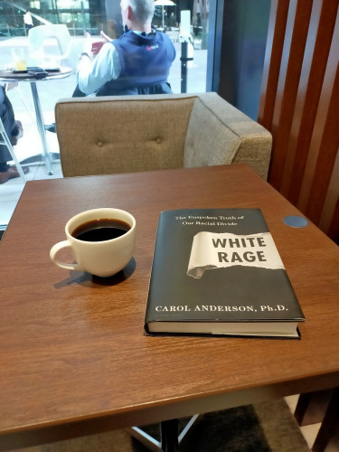 The photo is of a square brown wooden table inside the coffee house. The hardcover black book with a white scroll featuring the WHITE RAGE title in black lettering. To the left is a white mug of black coffee. A grey sectional chair can be seen across the table. Out the window can be seen an older short grey haired white man in a blue vest & white dress shirt sitting at a silver metal table outside in the day time