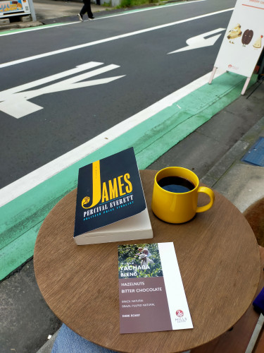 Photo of a small round table outside a cafe along a street with a long green line bordered by a white line. In the distance we can see someone's feet. Closer we can see white signboard on the right. On the table is a black paperback book with the word JAMES in yellow with the J extending to the top of the cover. To the right is a mug of black coffee. The mug is of the same yellow color. In front is a brochure explaining the coffee - YACHABA BLEND; HAZELNUTS, BITTER CHOCOLATE; BRAZIL NATURAL, BRAZIL PULPED NATURAL; DARK ROAST; MILLS COFFEE with a photo of 3 men in a coffee field with white hats and towels around their necks