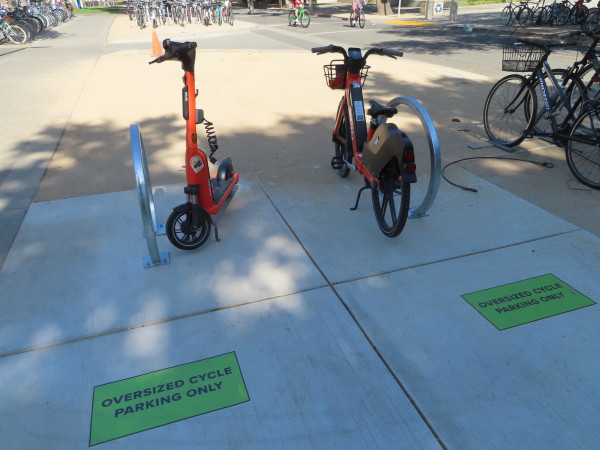 A SPIN electric kick scooter and a SPIN electric bike are both parked at the more accessible loop cycle parking on UC Davis campus shown upthread despite signage that makes it obvious who it's reserved for. Nearby are inaccessible "lightning bolt" racks.