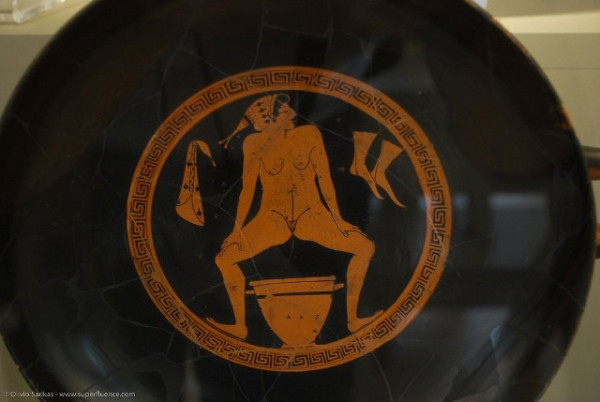 Red-figure vase painting of a woman peeing into a bucket. She is depicted fully nude from the front with a pubic triangle of either stubble or hair.