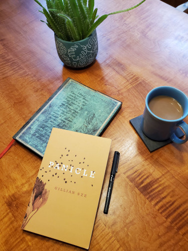 Poetry collection Panicle by Gillian Sze (ECW Press), with its subtle gold-toned cover and image of plants and seeds, sits on a wooden dining room table with a notebook with an ornate green cover, a succulent plant in a small teal and white pot, coffee in a blue cup and a black pen