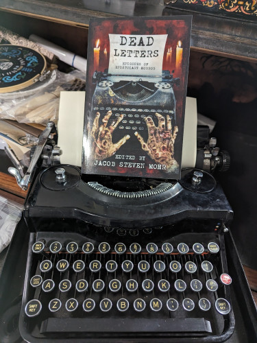 Paperback of DEAD LETTERS which has two decaying hands typing in an antique/vintage typewriter. I put the book on my antique/vintage typewriter for fun.