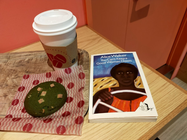 The photo is of a wooden counter on which is the paperback book which has a painting of a short haired Black woman in a car  looking forward with her right hand on a huge white steering wheel. She is wearing red spaghetti strapped dress. Behind her is the car's leopard print seat and blue behind that. To the left is a brown tray on which is a white coffee cup with a white plastic lid. It is brown sleeve with 3 big red beans. In front of it is a green Cookie with white chocolate chunks. It is on a red patterned paper sheet that says COSTA in round red circles