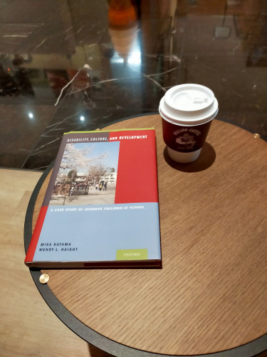 Photo of an indoor round brown cafe table. There is a coffee cup at the center top. It has a white plastic lid and a brown sleeve where the logo can fuzzily be seen. To the left is the hardcover book of modernist red and blue rectangles with a photo on the left of an elementary school with blurry Japanese students visible along with a pink cherry blossom tree