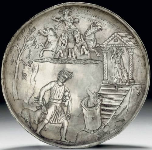 Silver bowl with a sacrifice scene in relief: a youthful priest wearing a flowing sleeveless tunic and a mantle rolled around his waist and draped over his left arm, a laurel wreath in his full curly locks, cradling pomegranates and other fruit in the crook of his left arm, gripping a goat by its horn, leading it toward a flaming hexagonal altar, embellished with scrolling dots on the upper surface and base, a wreath around its center, the altar situated at the base of the steps of a Temple to Athena, based on the Athena Parthenos. Helios driving His quadriga above, emerging from the clouds, the horses rearing, with two facing out to the sides, two facing in, the God with the right arm raised, an orb in the lowered right hand, a large tree along the left side, a Greek inscription to the right of Helios, reading: “Nonnos, priest, worshipper of Pallas, made an offering”.