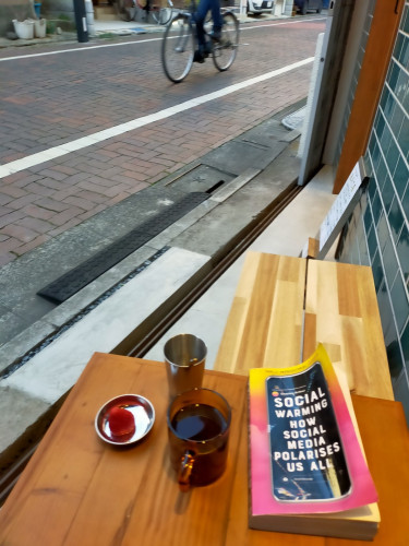 Photo of a wooden bench and table outside the cafe. The table's wood is darker than the light wooden bench  On the table is the paperback book which is yellow blending to orange blending to pink top to bottom. In the middle is black cracked screen smartphone with the title. To the left is a clear brown coffee cup of black coffee. Behind it is a silver cylinder cup. To the mug's left is a tiny round silver plate with a red strawberry in it. In the distance on the brick alley is a bicycle pedaling to the left. The rider's torso is cut off.