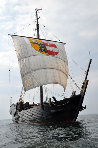 The replica cog "Wissemara" of Wismar. A dark wooden ship with one mast, bearing one large, white sail with coat of arms.