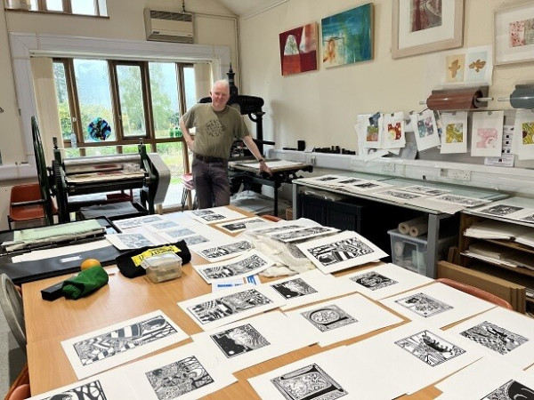 The printmaker in his studio, with completed prints spread out on the table in the foreground, and the presses in the background.