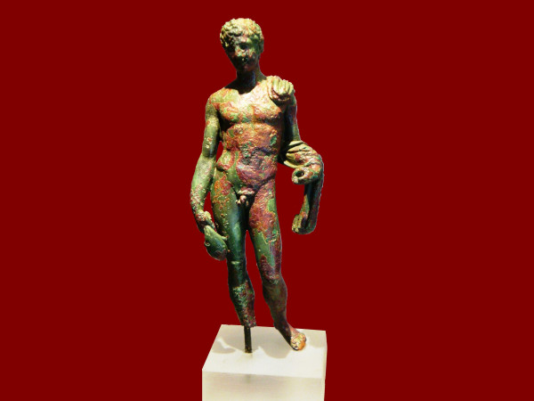 A bronze statuette of Hermes against a red background. The god is depicted in the nude with short-cropped, curly hair. A piece of fabric is slung over his left shoulder and draped around his left arm. He is holding a money purse in his right hand, arm hanging down relaxed by his side. His left hand used to hold his kerykeion staff but the staff was lost.