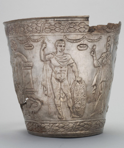 Silver bucket depicting Ares-Mars in the nude, wearing just boots and a cloak around his shoulders that is pinned over his right shoulder and covers only his chest. His hair is short and he is holding his spear in his right and a decorated shield in his left. Aphrodite stands to his left, fully clothed, holding her signature apple.