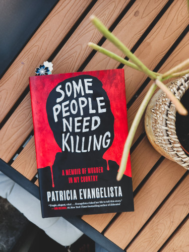The book titled Some People Need Killing by Partricia Evangelista on a slatted table. Also seen is a potted pencis cactus.