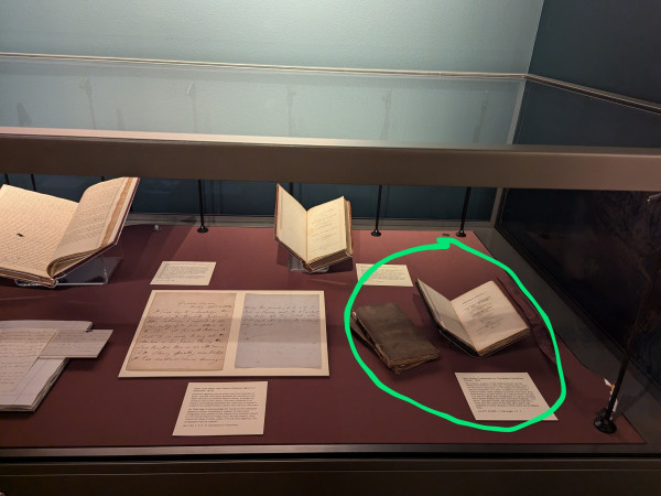 Display case with maroon fabric base. At right, volume open on a cradle. To the immediate left, two closed volumes, one stop another.

Rest of case in picture includes two volumes displayed open and a two page letter.