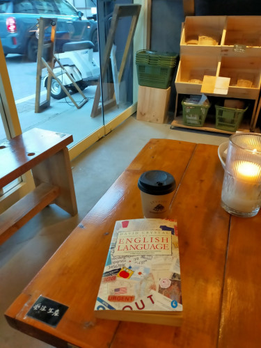 Photo is of a inside wooden table. The paperback book is on the table featuring a collage of travel items such as train stamps and tickets  Above the book is a brown coffee cup with a black lid. You can see a car passing upwards through the window of the door. You can see green carryable plastic baskets and grocery items & craft beers against the wall. A white candle is lit inside a corrugated glass lantern on the table to the left.