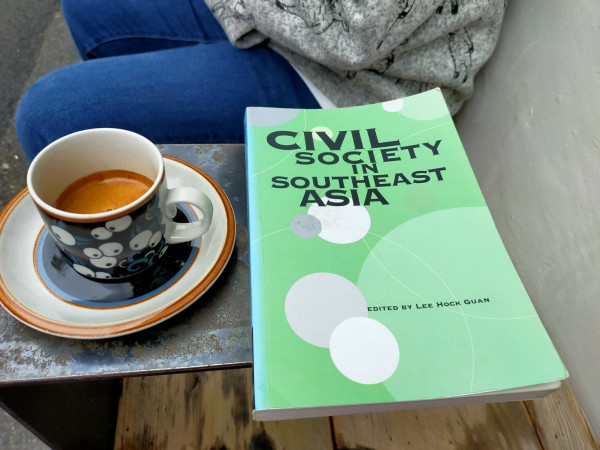 The photo is of a small metal table above a wooden bench. The paperback book mentioned in the toot is mint green with some white, grey, & lighter green circles of various sizes. To the left is an espresso cup with the crema visible. The copy has many white eyeballs among a blue background. the saucer is a white ring around a blue ring. A woman's blue jeaned legs in a light grey jacket with standing sheep, but not her face, can be seen behind the table.