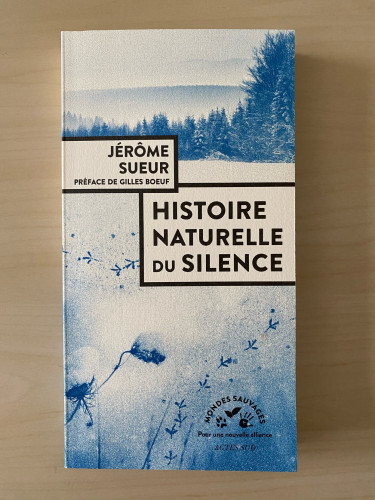 A book on a light brown table. The book is white and blue. The title Histoire Naturelle du Silence is written in large black letters in a white rectangle. The cover image shows a mountain and trees in the background, and what seems to be traces of birds in the foreground.
