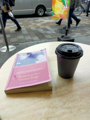 The photo is of an outside cafe table on which is a pink paperback book with a b/w photo of a white man in a wedding suit on the left with his back against an Asian woman with her black hair up smiling. To the right is a black paper coffee cup and black lid. In the distance you can see legs of people walking on the sidewalk & a car further beyond