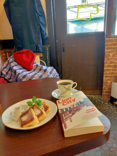 The photo is of a table inside of a cafe. On the table is the paperback book with an illustration of old Singapore underneath modern Singapore. To the left is a plate of french toast. Above the book is a cup & saucer of black coffee with a flower design. A dark & light blue & purple inside of a purple jacket can be seen on the chair across. You can see a wooden door further in the distance.