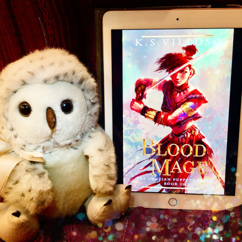 A photo of Sylvia, our spokes-owl, posing with the cover of Blood Mage by K.S. Villoso