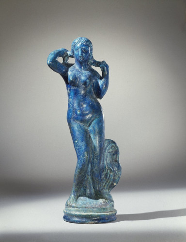 The statuette combines a classical image of the Greek goddess Aphrodite as she emerges from the sea, with the typically Egyptian material, faience. The goddess is shown entirely nude, her hands raised to wring out her hair. At her left is a greatly schematized urn with garment thrown over it. She stands on a round base with mouldings.