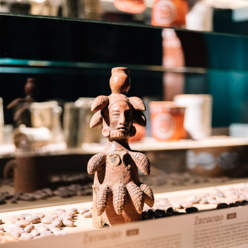 A small figurine of the Mayan goddess Ixcacao, the goddess of cocoa. She is wearing a skirt and hat decorated with cocoa pods and she is photographed standing among real life cocoa beans.