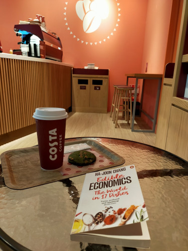 The photo is of the inside of the cafe. The white paperback book is on a round glass table. Above it is a tray on which is a red paper coffee cup with "Costa Coffee" vertical in white and "Costa" in a larger font  To tye right is a green matcha cookie with white chocolate squares. In the distance we can see the red espresso machine on a slatted wood counter on the left. On the wall directly across is a red wall with 3 large coffee beans. Below it is a garbage can for plastic & paper separate. To the right are wooden stools against a red counter.