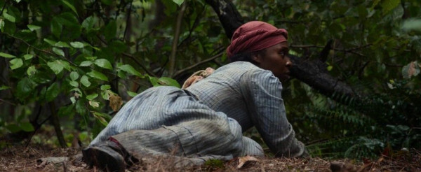 A young Black woman rises from the ground in the woods where she's been sleeping. She's wearing a dress and head scarf. She woke slowly, but now a sound has startled her, and she prepares to jump up