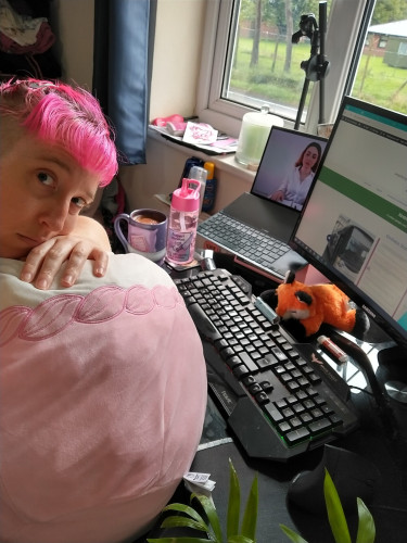 Alexis, a woman with pink hair in a fringe and an undercut, leans on a squishmallow which is on her lap while sitting at a desk. A laptop plays ASMR in the background and she has a bottle of water and a mocha in a large mug on the desk.