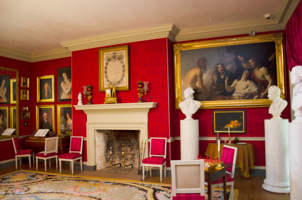 A lavish parlor room in the Madisons' home. The room features a red flocked wallpaper, French carpet, red upholstered French chairs, gilded framed paintings, marble busts on pedestals, a piano, a gilded clock, argand lamps, and a copy of the Declaration of Independence framed over the mantel. 