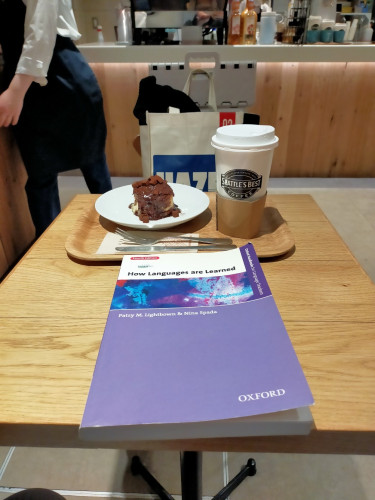 The photo is inside a cafe. The purple paperback book is in front on the table. Behind it is a light brown tray on which is a chocolate cinnamon roll on a white plate with a perpendicular fork & knife in front on a napkin. To the right is a white paper coffee cup with a white plastic lid and a brown coffee sleeve at the bottom. The cup has the Seattle's Best Coffee logo. A barista in a black apron is to the left but you cannot see her face.