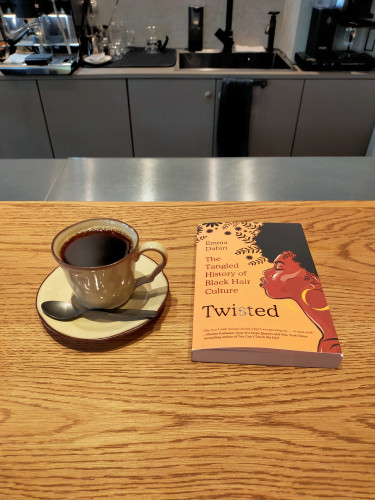 The photo is of a light wooden grained counter inside the coffee shop. In the distance are steel cabinets with a sink and various coffee making implements on top. In the foreground is the orange paperback book with a profile illustration of a proud Black woman with a natural afro and a big gold loop earring. To the left is a grey saucer & mug of black coffee with a tiny silver spoon in front perpendicular facing left