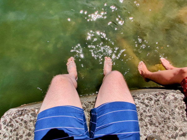 The photographer's pasty white legs dangling in the water from a concrete pier. The dark skinned feet of the photographer's wife (with light green painted toenails) also dangle in the water on the right.