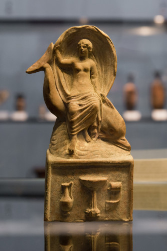 Aphrodite riding a dolphin over the sea. The sweeping movement of the cloth, far over the head of the goddess, represents the speed of the movement. The figurine is atop what looks like an altar.