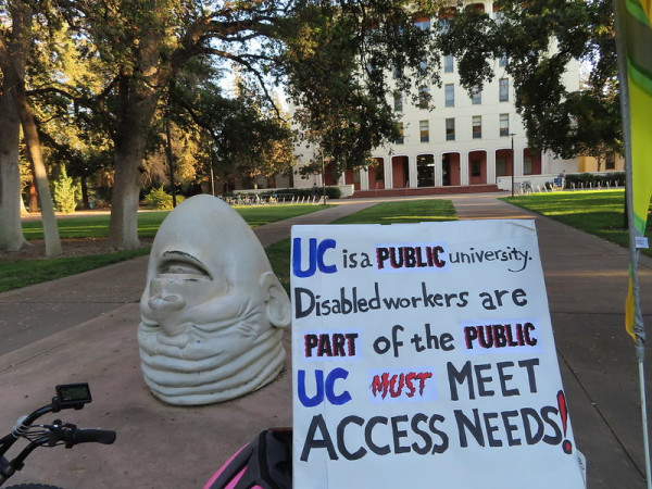 Laughing Egghead sculpture in front of the UC Davis Administration Building - Mrak Hall. The quadricycle is parked in front of it with a strike rally sign that says "UC is a public university. Disabled workers are part of the public. UC must meet access needs!"