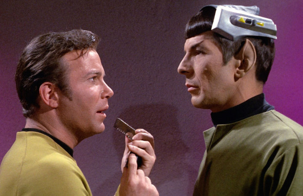 Kirk speaking into his tricorder with Spock facing him and wearing a mechanical contraption on his head