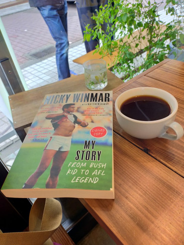 The photo is of the corner of a square brown table. The paperback book featuring the iconic photo of Aboriginal Australian Rules Footballer Nicky Winmar lifting up his St Kilda Saints jersey pointing proudly at his skin in a gesture of defiance at racial abuse in 1993. To the left is a white coffee mug of black coffee. 2 pairs of legs can be seen outside on the sidewalk through the window