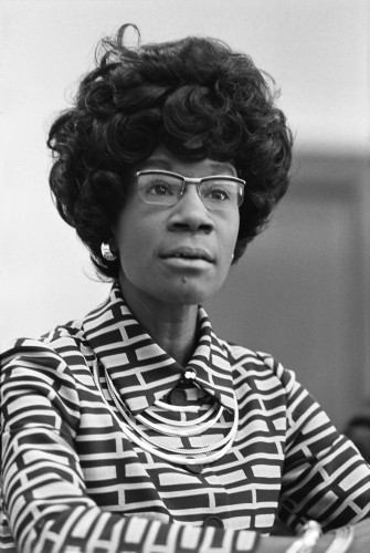 Black and white formal photo of Shirley Chisholm, the first Black woman elected to the House of Representatives. She has a 60's style hairstyle, not sure what to call it--maybe bee hive?  Glasses, and she is wearing a geometric patterned shirt with a buttoned collar and a necklace with multiple tiers.  