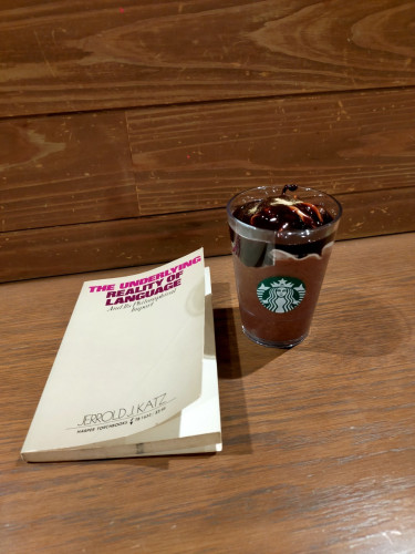 The photo is of a wooden counter on which is the grey paperback book & next to it is a glass of chocolate Opera frappuccino.  The glass has the green Starbucks mermaid logo in the center
