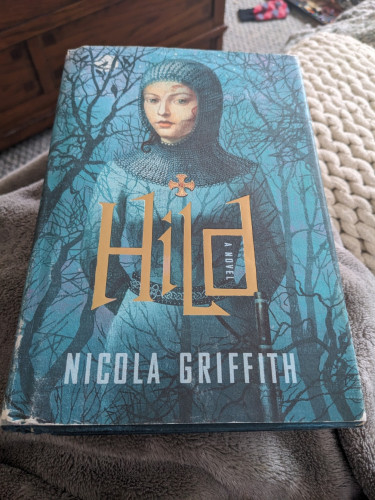 A hardcover copy of Hild by Nicola Griffith: a tall young woman stands wearing armor and a chainmail coif with wisps of reddish hair peeping out.  She wears a belt with an Anglo-Saxon motif and a large knife. She almost blends into a greenish blue background, which also has spindly, leafless trees.