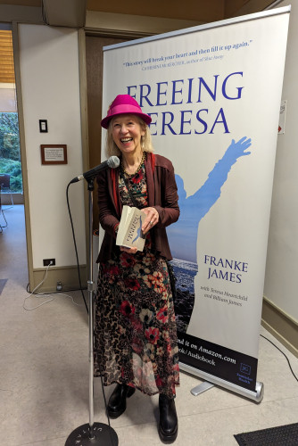 Author Franke James at the book launch for 'Freeing Teresa' at Spectrum Society in Vancouver. She's smiling and holding a copy of the new book. A microphone stands in front of her. Franke is wearing a magenta hat, a long floral dress, and black boots. She is in front of a large  'Freeing Teresa' book poster.