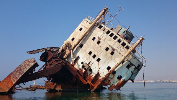 Photograph shows the remains of a large ocean vessel leaning as it rests in shallow water. The hull of the ship is rusted, tattered and demolished.  All that remains are rusted beams and scraps metal dangle from the upper deck's carcass.  The image is symbolizes the poor and disabled huddled masses, as well as the American political system. Time wasted on bickering and bureaucracy, has taken a toll in many ways.