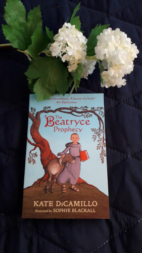 The cover of The Beatryce Prophecy shows a little girl, her hair cut to a buzz, wearing monk robes and holding a red book in one hand while petting a goat with the other.