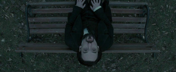 A dark-haired man tilts his head back on a bench, looking straight up into the sky