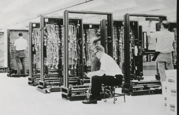 Assembly workers working on the wiring of the Control Data 3000 Series computers. Either standing or seated there is one worker to a frame of the machine working on wiring it.  Black and white horizontal oriented rectangular photograph from 1962.