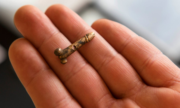 Photo of a human hand holding a tiny golden phallus with a hole to wear as a pendant. Two small testicles were fashioned at the base.